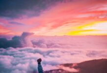 성취도를 높이는 방법 man sitting on mountain cliff facing white clouds rising one hand at golden hour