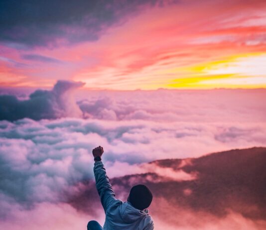 성취도를 높이는 방법 man sitting on mountain cliff facing white clouds rising one hand at golden hour