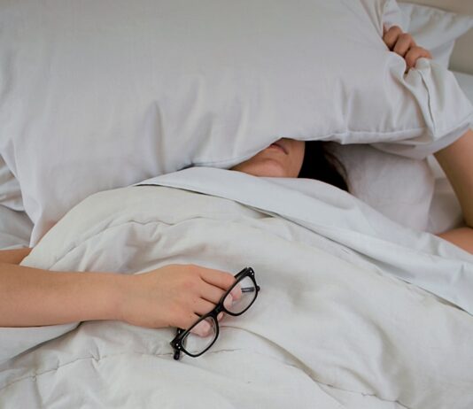 “밤에 자꾸 깨요”…수면장애의 뜻밖의 원인, ‘체온 조절 실패’ person lying on bed while covering face with pillow and holding eyeglasses